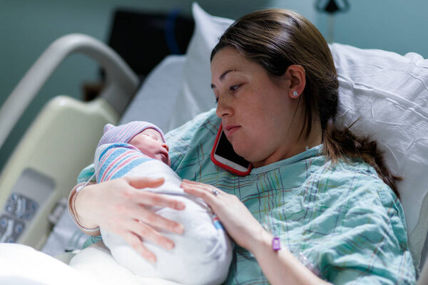 mother and her newborn baby in a hospital maternity ward room as mom talks on her cell phone
