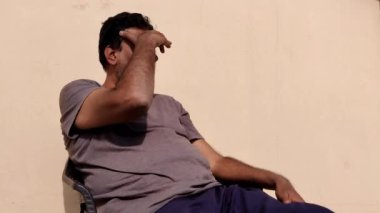 Güneşli bir günde, güneşli bir günde dışarıda oturan orta yaşlı bir adam gri tişörtü ve mavi pantolonuyla tek eliyle gözlerini güneş ışığından koruyan bir adam..