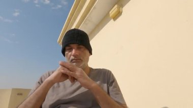 Stresli adam evinin önündeki terasta oturuyor mavi gökyüzünün altında ve parlayan güneşin altında