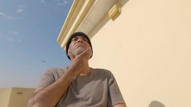 Adam küçük beyaz traşını eliyle ovuyor. Siyah bir şapka takıyor ve güneşli bir günde mavi gökyüzünün altında oturuyor.