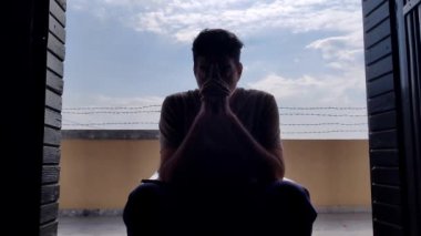 Kapının arasında oturan bir adam endişeli görünüyor, elleriyle burnunu ovuyor. Arkasında, gökyüzü mavi ve duvarda dikenli teller var..