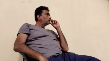 Bir adam sandalyede çok dağınık bir şekilde oturuyor, derin düşüncelere bakıyor. Saçları siyah ve huzursuz görünüyor..