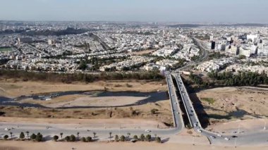 Hava görüntüsünde, her iki yönde de yol trafiği akan ve gündüz vakti görülebilen bir şehir manzarası bulunan kurumuş bir nehir yatağı üzerine inşa edilmiş bir köprü yer alıyor..