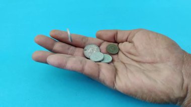 Bozukluklar yavaş çekimde açık bir avuç içine dökülür yatıştırıcı mavi bir arka plana karşı. Düşen paraların metalik parıltısı, ekonomik mücadelelerdeki mali risk, istikrarsızlık veya geçici fırsatlar temalarını çağrıştıran huzurlu arka planla çelişiyor.