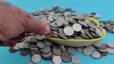 Bir kase açık mavi bir zemin üzerinde madeni paralarla dolup taşarken, bir el büyüyen yığının içine ilave madeni paralar koyar. Bu sahne gayretli tasarruf, finansal disiplin ve istikrarlı zenginlik birikimini simgeliyor., 