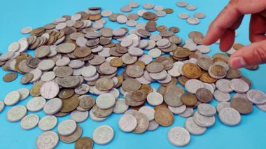 El bir yığın madeni paraya uzanıyor ve bir tanesini kaldırıyor, bu da yatırımların azaltılması veya maddi kaybın sembolize edilmesi anlamına geliyor. Açık mavi arka plan metalik madeni paralarla çelişiyor, risk dengesizliği temaları çağrıştırıyor ve ihtiyatlı mali planlama