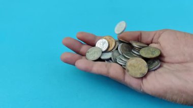 Bir el dağınık sikkeleri tutarken yukarıdan daha fazlası düşüyor, bazıları açık mavi arka planda avuç içinden kayıyor. Bu görsel metafor finansal dengesizliği, çabayı ve geçici zenginliği temsil eder.