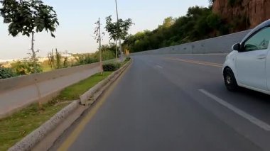 2 Nisan 2025 Rawalpindi Pakistan banliyösünde şehir yolu ve yeşil çimenli patika üzerinde hareket eden bir arabayı gösteren bisiklet kaydedilen video.