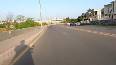 Yolda görünür gölgesi olan motosikletli bir adam var. Arka planda seyrek trafik var. Şehir içi ulaşımı gösteriyor.