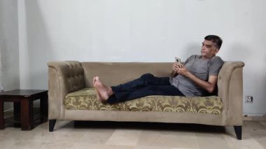 Koltukta bacaklarıyla oturan adam, özel espri anlayışını yansıtan telefona bakarken muzip bir şekilde gülümsüyor.