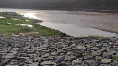 Parlak güneş ışığı altında akan nehir huzurlu bir manzarada sabit dağ taşlarının yanında ışığı yansıtıyor.