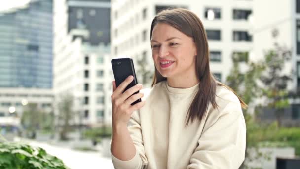 耳にイヤホンを装着した若い陽気な女性は 現代のヨーロッパの街の通りに座っている間 スマートフォンで話しています — ストック動画