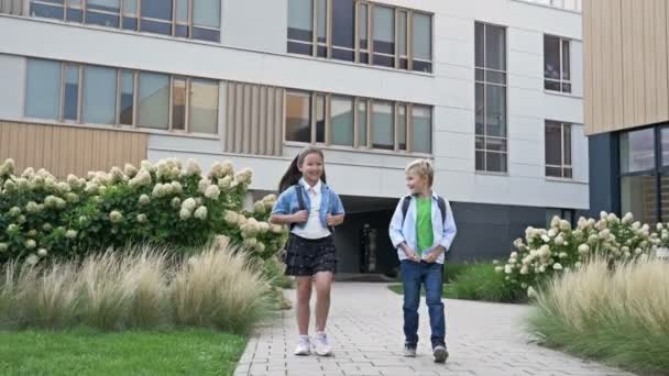 2人の小さなクラスメート 男の子と女の子は 良い気分で学校の庭を歩いている 学校の友情 — ストック動画