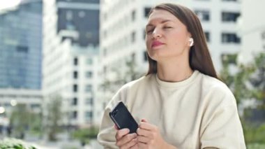 Akıllı telefon ve kablosuz kulaklık kullanan çekici bir kadın müzik dinliyor. Modern Avrupa şehri, yaz günü. Kadın zevkle gözlerini kapar ve dans eder. HD.