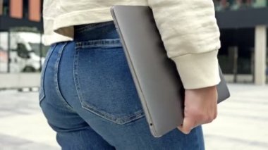Elinde dizüstü bilgisayarı olan kot pantolonlu bir kadın şehir caddesinde yürüyor. 4K.