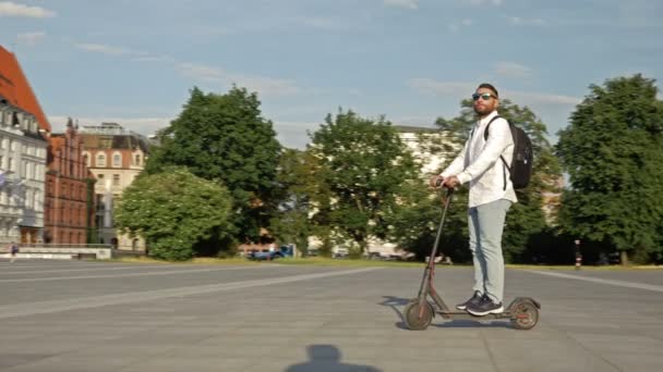 肩にリュックを背負った白いシャツの男が電動スクーターに乗って街の広場を通っています 便利で環境に優しい個々の都市交通の概念 — ストック動画