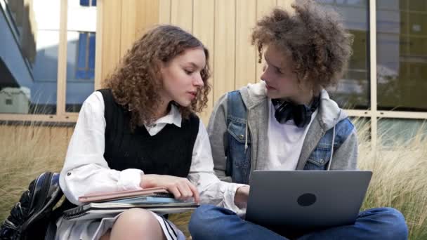 2人の可愛い高校生 男と女が校庭のベンチに腰を下ろしている 10代の若者はノートパソコンの画面を見て 何かを議論する楽しみがあります — ストック動画