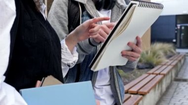 İki lise öğrencisi, bir erkek ve bir kız, bir bankta oturuyorlar ve bir şeyi hararetle tartışıyorlar. Adamın elinde büyük bir defter var. HD.