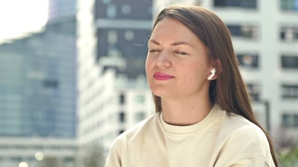 耳にワイヤレスヘッドフォンを持つ若い魅力的な女性は音楽を聴く 現代ヨーロッパの都市 夏の日 女は喜んで目を閉じる — ストック動画