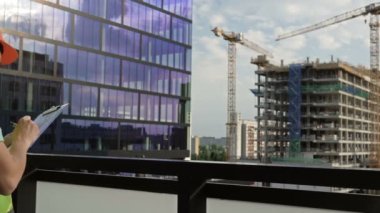 Young female builder in a signal vest and helmet writes something. Against the backdrop of a building under construction. HD.