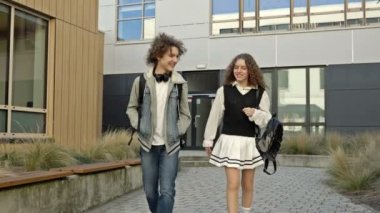 İki sevimli lise öğrencisi, bir erkek ve bir kız, eğitim kurumunun avlusunda dolaşıyorlar. Gençler hareketli konuşuyorlar. 4K.