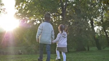 İki küçük çocuk, bir büyük oğlan ve bir küçük kız, parkta el ele tutuşarak yürüyorlar. Gün batımı. Dikiz aynası. 4K.