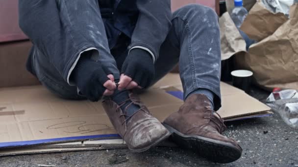 ゴミの山に座っているホームレスの男は震え 汚れた手で彼のテータリングブーツを結ぶしようとします — ストック動画