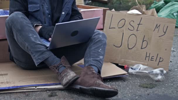人在寒冷中颤抖 膝盖上架着笔记本电脑 坐在垃圾堆中的纸板上 旁边是一张手写的海报 上面写着 失去我的工作 失业信息技术专家 — 图库视频影像