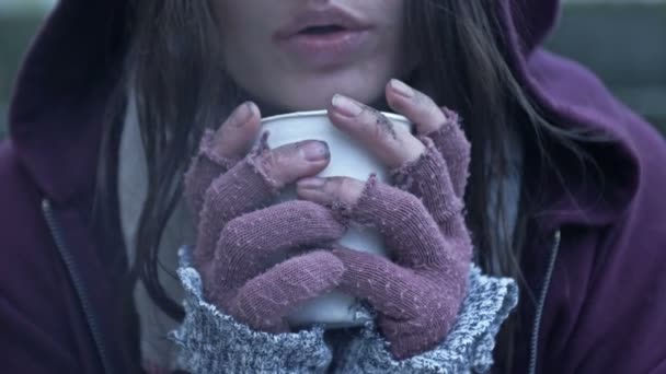 冷たいから震えているホームレスの若い女性は 汚れた手にある種の飲み物の紙コップを持っている — ストック動画