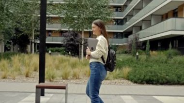 Sırt çantalı ve elinde dizüstü bilgisayarı olan neşeli genç bir kadın bir yaz şehrinin caddesinde hafifçe yürüyor. HD.