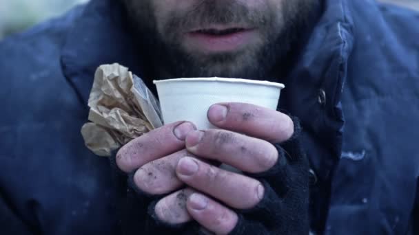 冷たい顔をしたホームレスの男は 汚れた手にある種の飲み物の紙コップを持っている — ストック動画