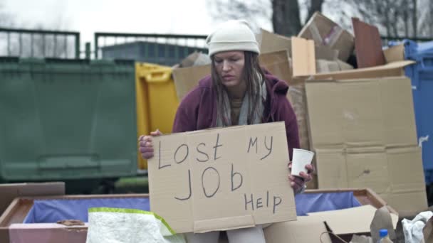 冷たい顔をしていない若い女性は ごみの山によって物乞いカップに座って 手書きの失われた私の仕事ヘルプポスターを保持しています — ストック動画
