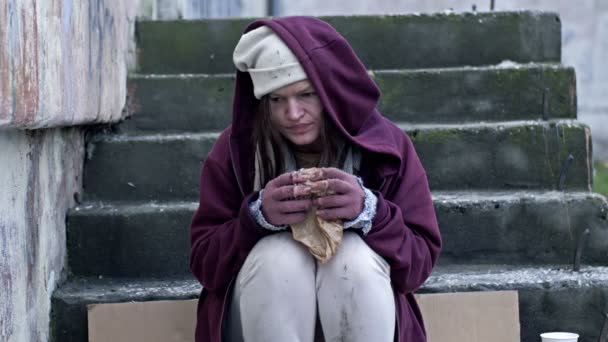 ホームレスの若い女性は 寒さに震え 貪欲に紙に包まれた何かを食べる 彼女は貧弱で汚れていて助けを必要としている — ストック動画