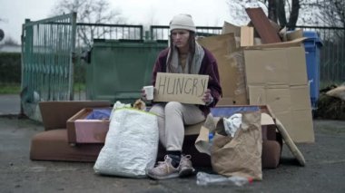 Donmuş, dağınık görünümlü genç bir kadın bir çöp yığınının yanında oturuyor ve elinde bir HUNGRY posteri tutuyor. Yoldan geçen biri dilenci bardağına birkaç bozuk para atar. 4K.