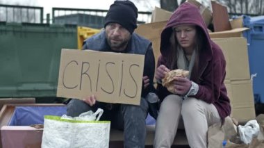 İki kirli ve kötü giyimli evsiz insan, bir adam ve bir kadın, ellerinde CRISIS posteri olan bir çöp yığınının yanında oturuyorlar. Çiftler bir güvercin sürüsü tarafından korkutulup alkışlanıyor. 4K.