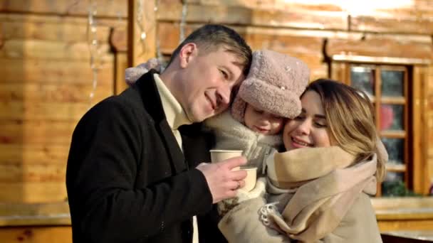 一对年轻漂亮的夫妇和他们的小女儿在圣诞集市上 父母温柔地把孩子抱在怀里 成年人手里拿着纸杯热饮 — 图库视频影像