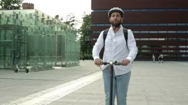 Sırt çantalı ve koruyucu kasklı sakallı bir adam şehir meydanında elektrikli scooter kullanıyor. Kullanışlı ve çevre dostu bireysel şehir taşımacılığı kavramı. HD.