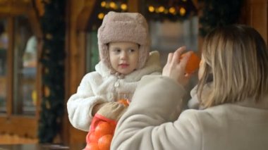 Anne küçük kızını eğlendirir. Kadın gözlerine mandalina sürüyor, çocuk eğleniyor. Bir Noel fuarı. HD.