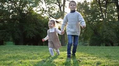 İki küçük çocuk, bir büyük oğlan ve bir küçük kız, parkta el ele tutuşarak yürüyorlar. Çocukların keyfi yerinde ve birbirlerine bakarken gülüyorlar. HD.