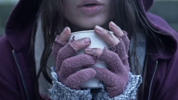 冷たいから震えているホームレスの若い女性は 汚れた手にある種の飲み物の紙コップを持っている — ストック動画