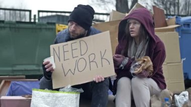 Two sad homeless men sit by the rubbish bins. Man and a woman are poor and dirty dressed. The man is holding a begging glass and a NEED WORK poster. Young woman is sick and coughing. HD.