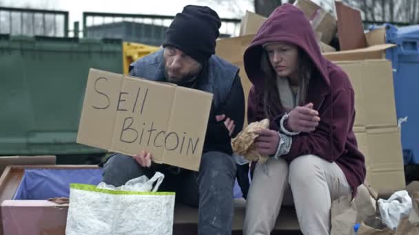 汚い服を着た2人のホームレスの人々 男性と女性は 手書きのSell Bitcoinポスターでゴミの山に座っています 若い女性は病気で咳をしている — ストック動画