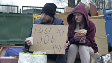 Two dirty and poorly dressed homeless people, a man and a woman, sit by a pile of rubbish with a handwritten LAST MY JOB HELP poster. Passer-by tosses a few coins into a begging cup. 4K.