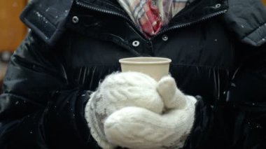 Winter portrait of a woman in warm clothing warming herself with a hot drink. Light snow is falling. 4K.
