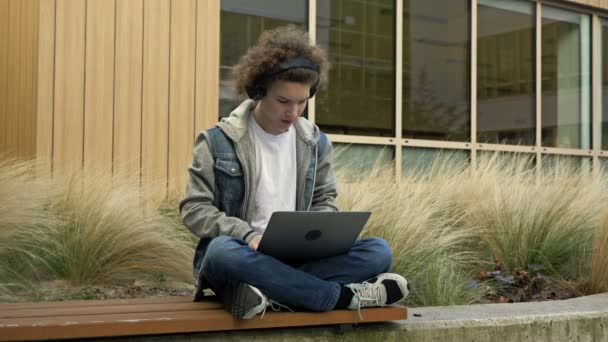 教育機関の庭にあるベンチにラップトップで座っている男は何かに圧倒されています コンピュータゲーム 良いニュース 証券取引所での幸運を獲得 — ストック動画