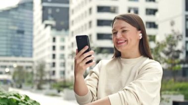 Young cheerful woman with an earphone in her ear is talking on a smartphone while sitting on the street of a modern European city. 4K.
