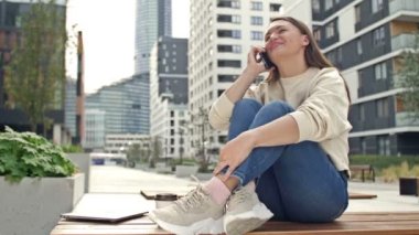 Young cheerful woman talking on a smartphone while sitting on the street of a modern European city. HD.