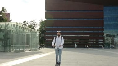 Sırt çantalı ve kasklı bir adam şehir meydanında elektrikli scooter kullanıyor. Kullanışlı ve çevre dostu bireysel şehir taşımacılığı kavramı. HD.
