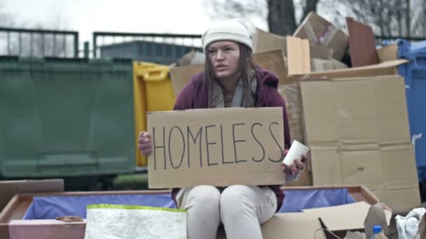 冷たい顔をしていない若い女性は ごみの山にゴボゴボというカップに座って手書きのホームレスのポスターを持っています — ストック動画