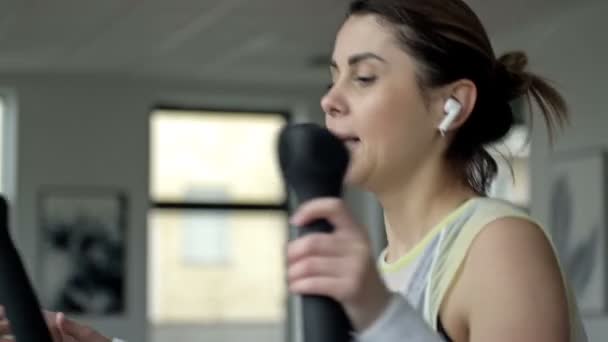 Kulaklıklı Genç Güzel Bir Kadın Spor Salonunda Bir Simülatörle Meşgul — Stok video